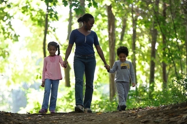 A mother stands between her two children holding their hands, walking through a forest. To the right, her daughter wears a pink shirt and jeans. The mother is in a blue top, and the son to the right has a tractor shirt on.