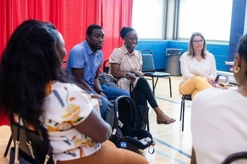 A small group of people sit in chairs arranged in a circle in a gym, engaged in a discussion. A red curtain backdrop and large windows are visible. A baby carrier rests on the floor beside one participant.