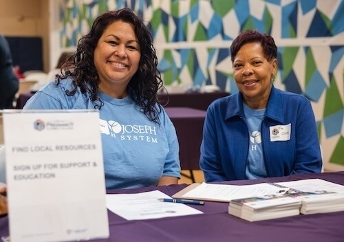 Two women smile at the camera from behind a table with a sign reading "Find Local Resources & Sign Up for Support & Education." They wear matching blue shirts with the words "Joseph Health System." Pamphlets and paper are arranged on the table.  A geometric teal, green, and white wall is visible in the background.