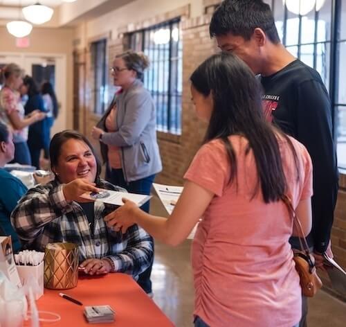 A woman sitting at a table hands informational pamphlets across an orange tablecloth to a pregnant woman and a man standing together. Other attendees and exposed brick walls are visible in the background of this bustling event.