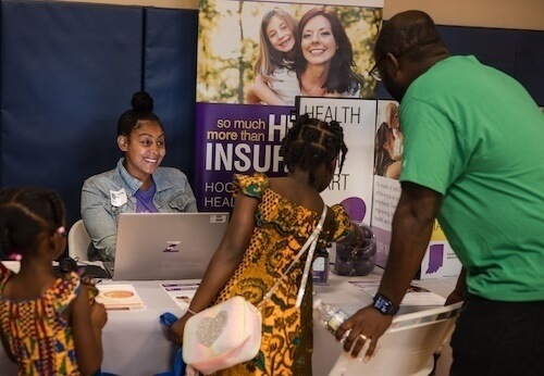 A woman sits at a table with a laptop, assisting a family at a health insurance information booth. Two children, dressed in vibrant patterned clothing, stand by the table while a man looks on. A banner behind the woman promotes health resources.