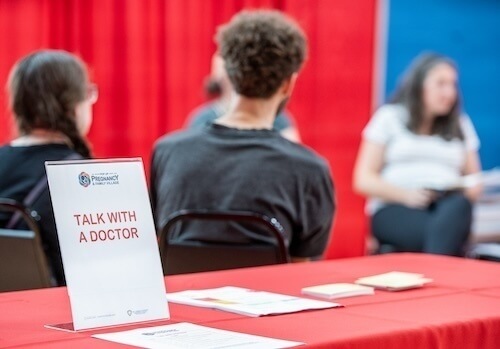 A sign reading "Talk With A Doctor" sits on a table covered with a red tablecloth in the foreground. In the background, several individuals sit in chairs with a red and blue backdrop. The image suggests a health information event, possibly related to the University of Notre Dame's Pregnancy and Family Village logo on the sign.