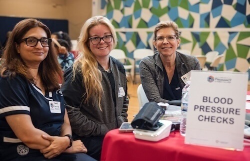 Three women sit at a table with blood pressure equipment and a sign reading "Blood Pressure Checks." They appear to be healthcare providers at a health fair.
