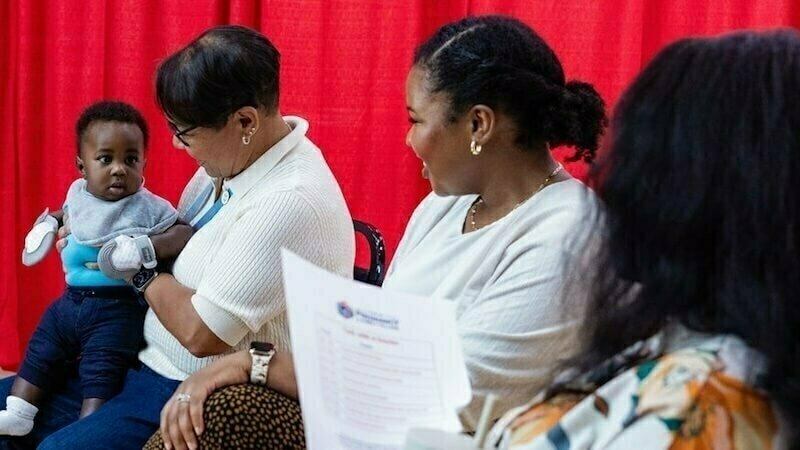 A woman holds a baby wearing a gray bib and gray mittens against a red backdrop. Two other women sit next to them, one looking at the baby and the other facing away. A partially visible document with the words &quot;Parental Consent&quot; is in the foreground.