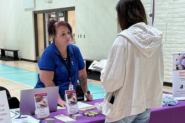A woman in a blue shirt speaks with a person wearing a light gray hoodie at an informational table with pamphlets and a laptop.