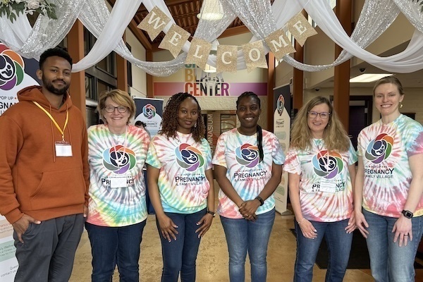 Six individuals wearing tie-dye shirts promoting Pregnancy & Family Village stand inside the Compton Family Ice Arena's LaBar Practice Center beneath a "Welcome" banner.