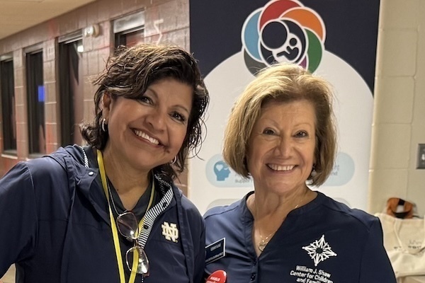 Two women smile in front of a banner for the William J. Shaw Center for Children and Families. One woman wears a navy Notre Dame jacket, and the other wears a navy William J. Shaw Center shirt.
