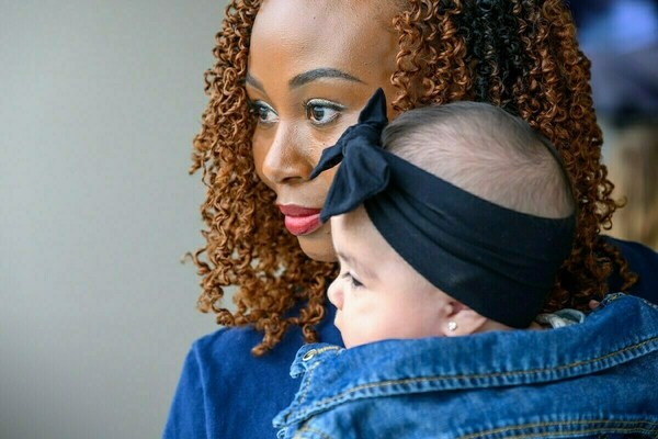 A woman with warm brown curly hair and red lipstick holds an infant wearing a black bow headband and denim jacket. Both gaze intently to the left.